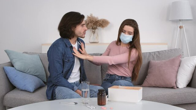 Young man trying to kiss his girlfriend, sick woman in protective mask stopping him, sitting on couch at home