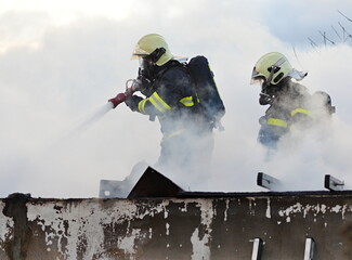 Firefighters with breathing apparatus masks extinguish the burning roof of a family house using...