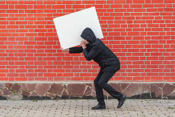 A man carries on his shoulder a blank white canvas for painting against a red brick wall 