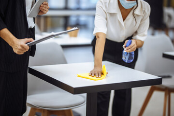 Young businesswomen clean the work place, wipe the desk with yellow rag. Colleagues disinfect the working surface with a sanitizer spray to stop covid-19 spread. New normal, office hygiene concept.