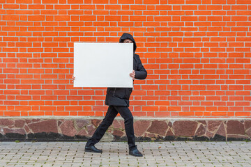 A man in black clothes moves along a red brick wall with a white blank canvas in his hands