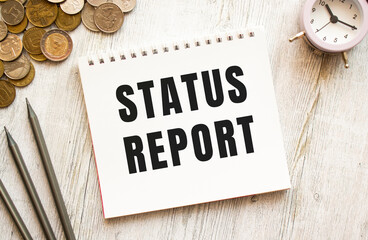 STATUS REPORT text on a sheet of notepad. Coins are scattered, pencils on a gray wooden background.