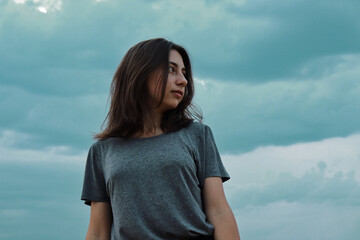 A cute girl with dark blond hair in a gray T-shirt looks to the side in a field against a blue sky on a summer day.