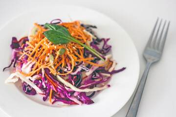 Vitamin vegan salad with red cabbage, carrots, olive oil and sesame seeds on a white plate with a fork on a white table. A healthy plant-based food concept made from local, seasonal ingredients.