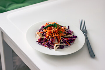 Vitamin vegan salad with red cabbage, carrots, olive oil and sesame seeds on a white plate with a fork on a white table. A healthy plant-based food concept made from local, seasonal ingredients.