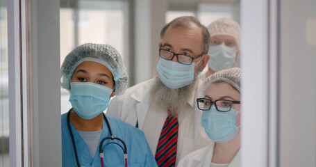 Team of healthcare workers in safety mask standing behind glass door watching difficult surgery
