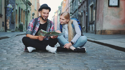 Young tourists couple using map, sitting on pavement and admiring historical surroundings. They are excited and smiled.