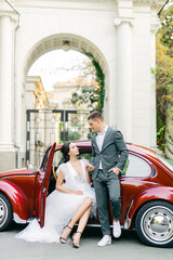 Fototapeta na wymiar Portrait of a happy young wife and husband hugging on the background of a red retro car in tropic garden. Sincere feelings of two young people. The concept of true immortal love. Elopement