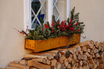 Window decoration for Christmas in the form of plant branches and red berries in a wooden pot on the building window. Folded neatly chopped firewood for barbecue fire