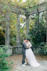 attractive bride in a delicate fashionable wedding dress kisses the groom in a gray suit near stone columns in a tropical garden on a warm summer day.
