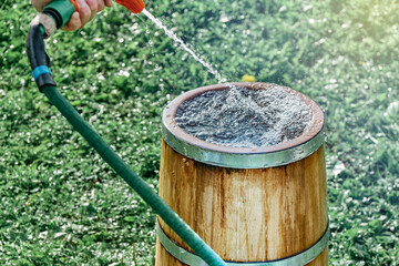 Man cleans or fills vintage oak wood tub with clear water from hose on lush green lawn grass on sunny summer day close view