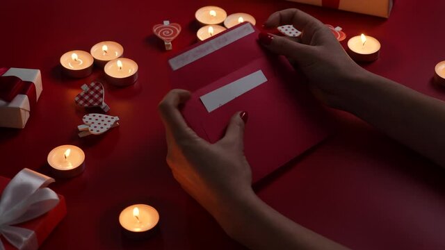 Woman's hands open envelope and take out a valentine card with inscription love you. Top view of red table with burning candles, decorative hearts and gift. Valentine's Day. Close up. Slow motion.