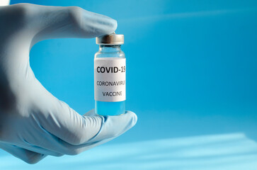 Hand in medical blue glove holding bottle of Covid 19 coronavirus vaccine on blue background with copy space and with shadows. Vaccination concept, inscription Covid 19 coronavirus Vaccine .