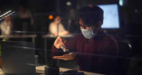 Asian businessman working in office at night wearing face mask, sitting at desk using hand sanitizer.
