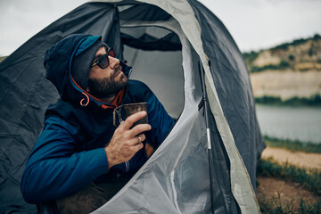 Man sitting in the tent, holding cup of coffee and looking at the sky.