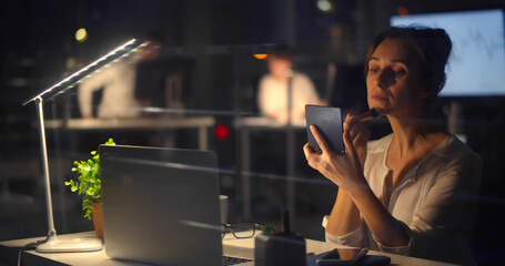 Young businesswoman doing makeup at workplace in dark office at night
