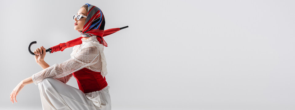 stylish woman in headscarf and sunglasses holding red umbrella while sitting isolated on white, banner