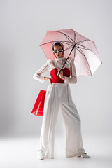 full length of stylish woman in headscarf and sunglasses holding red umbrella and shopping bag while posing on white