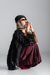 Naklejka premium full length of young woman in trendy outfit sitting and posing on grey