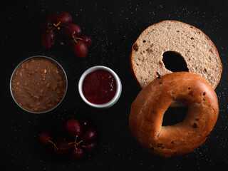 Raisin Bagel with pot of jam and crunchy peanut butter and red grapes on black surface with sprinkles of white sugar