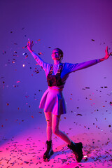 Fototapeta na wymiar full length of happy woman in stylish outfit with outstretched hands near falling confetti on purple