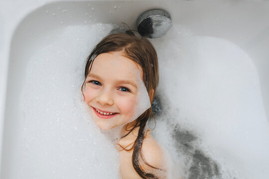 A small, smiling, cute red-haired girl with long hair, the child bathes, lying in a white bath with foam from soap, shampoo and indulges. Model photography.