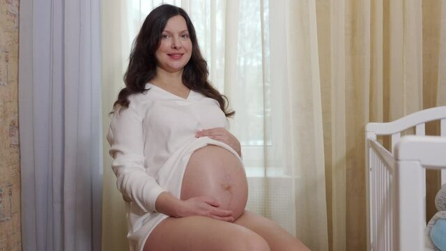 happy expecting mother sitting near white baby cot pregnant woman stroking belly. High quality 4k footage