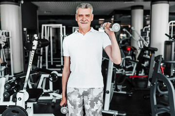 Portrait of a senior man in the gym with dumbbells in his hands, he performs an exercise and looks...