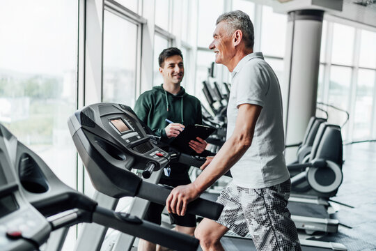 Portrait Of An Older Man On The Treadmill, With His Coach Standing Next To Him And Watching And Recording The Results. Rehabilitation. Training Plan, Coach And Patient