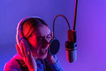 woman in wireless headphones recording song while singing in microphone on purple