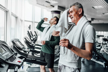 In focus, a senior man in the gym after training drinks water and wipes with a towel, on the back...