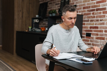 Fototapeta premium Serious man in earphones writing down notes and working with laptop