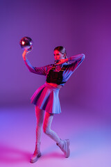 full length of trendy woman in skirt holding disco balls on purple
