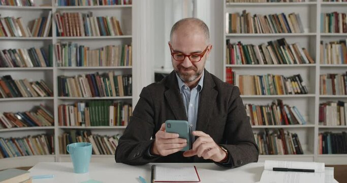 Crop view of bearded male person in suit using smartphone and smiling while sitting at desk. Mature man in glasses touching phone screen while chatting and texting in room full of books