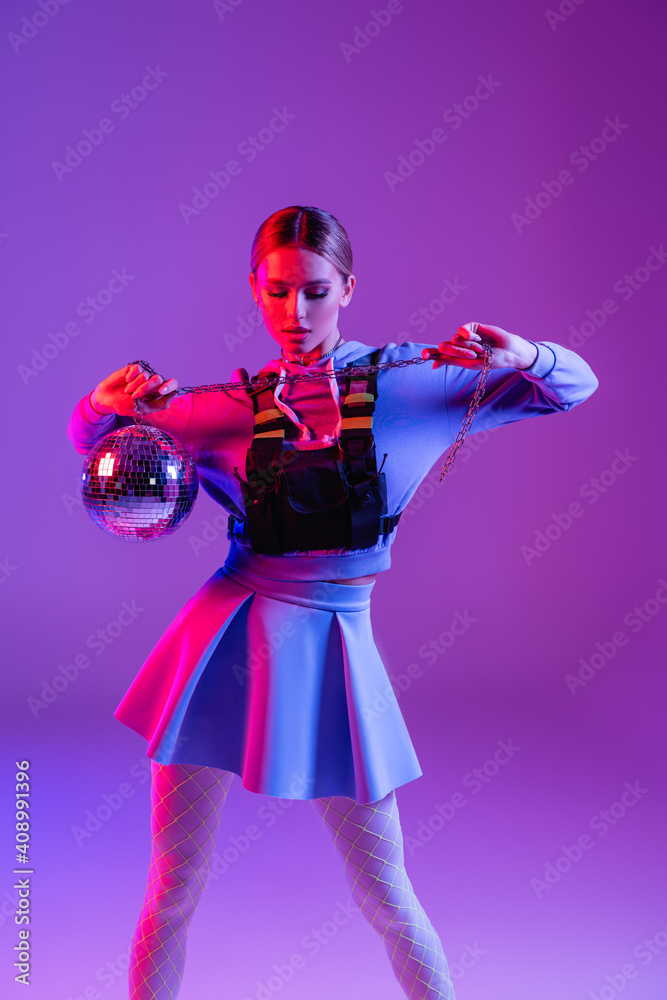 Poster young and stylish woman in skirt posing with shiny disco ball on purple