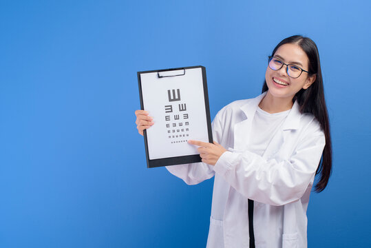 Young female ophthalmologist with glasses holding eye chart over blue background studio, healthcare concept