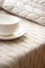 Fototapeta na wymiar Large white ceramic cup on a saucer on a knitted nude blanket. Cozy home background. Still life from home interior