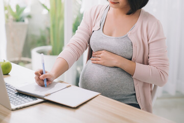 Closeup of a pregnant woman writing notes and using a laptop while working on maternity leave at her dining room table at home - 408996501