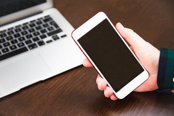 Close-up of a man's hand holding a smartphone and a laptop in the background. Concept of wireless technology and use of smartphone and laptop