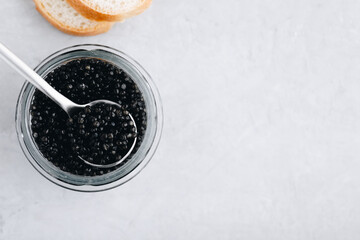 Black caviar in glass jar with spoon on white stone background, luxurious delicacy appetizer. Top view, copy space