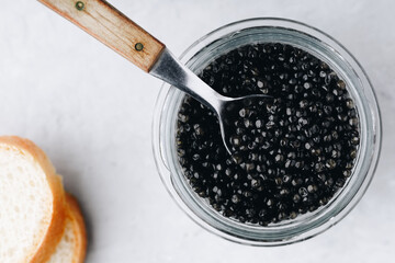 Black caviar in glass jar with spoon on white stone background, luxurious delicacy appetizer. Top view, copy space