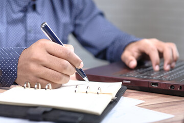 man's hand writing on notepad and working on laptop 