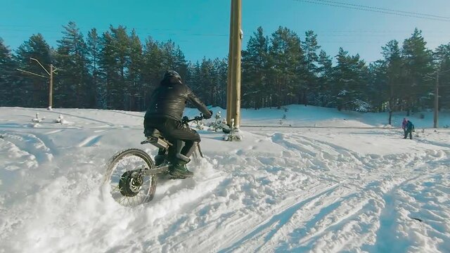 Cyclist in Black Clothes, Helmet and Snow Glasses Riding on Electric Bike in the Snow in Winter Forest. E-Bike Riding and Drifting at Cold Sunny Day - Super Slow Motion