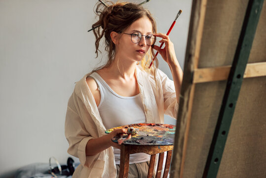 Horizontal candid shot of a girl artist standing next to the easel with canvas painting something in her art studio. A professional young woman painter in transparent eyeglasses draws in the workshop.