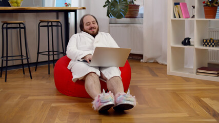Funny fat guy wearing bathrobe and unicorn slippers sitting in beanbag and using laptop