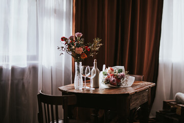 Glass vase with flowers on a wooden table in front of a light background. On the table are a bottle and glass wine glasses. Romantic mood for Valentine's day, Woman day, gift 8 march.
