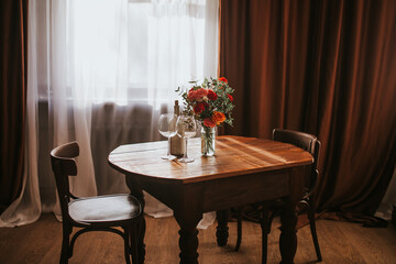 Glass vase with flowers on a wooden table in front of a light background. On the table are a bottle and glass wine glasses. Romantic mood for Valentine's day, Woman day, gift 8 march