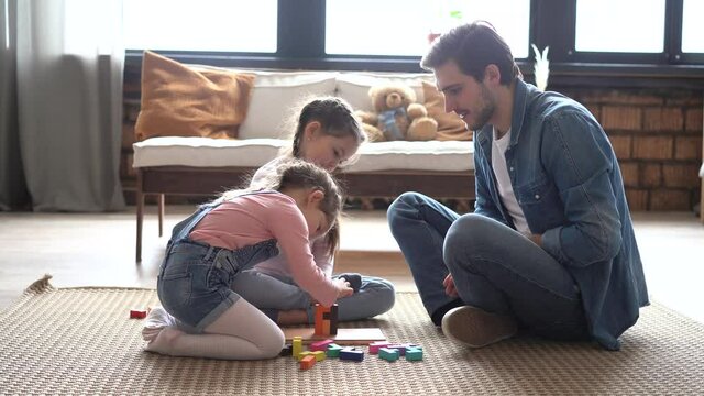 Happy parent dad helping daughters playing toys together on floor, having fun