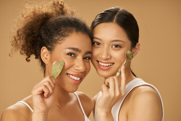 Cute young women smiling at camera, using jade roller and facial gua sha while posing together isolated over beige background