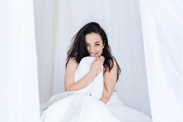 Happy young beautiful woman hiding under the blanket in her bed early in the morning